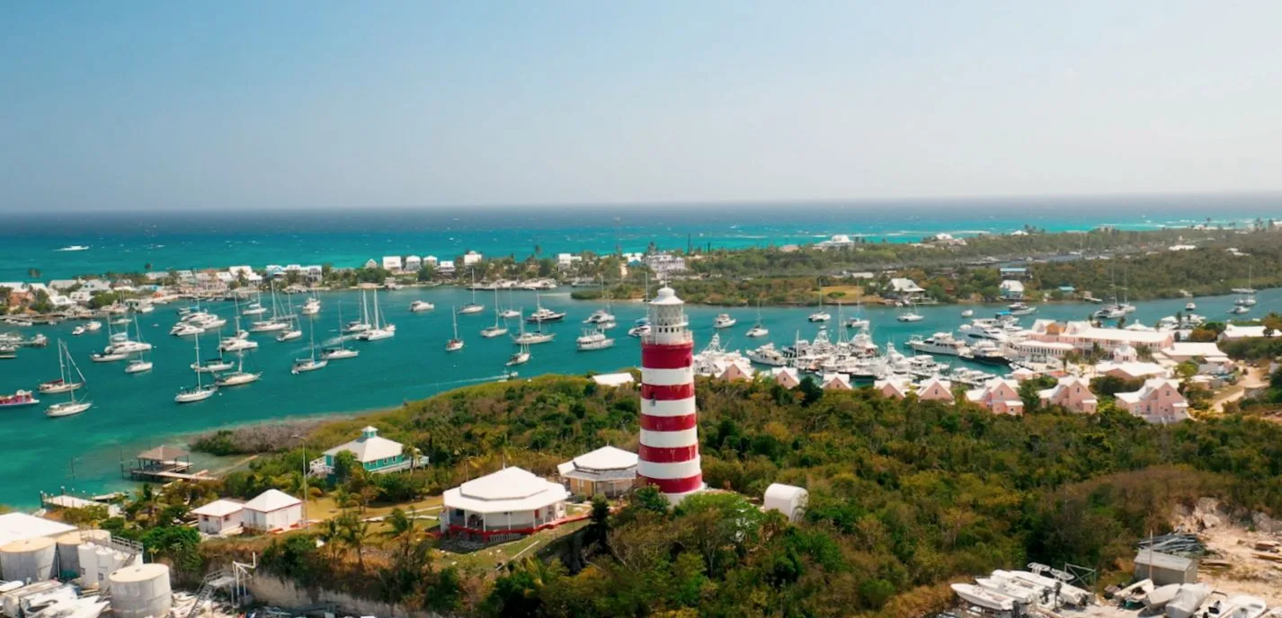 Aerial view of Hope Town, Elbow Cay — the red-and-white lighthouse rising over the harbour, catamarans at anchor, turquoise Sea of Abaco beyond